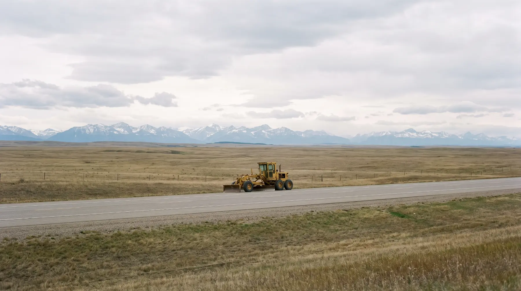 A motor grader parked on a highway shoulder with the prairie running out to the Rocky Mountains under an overcast sky